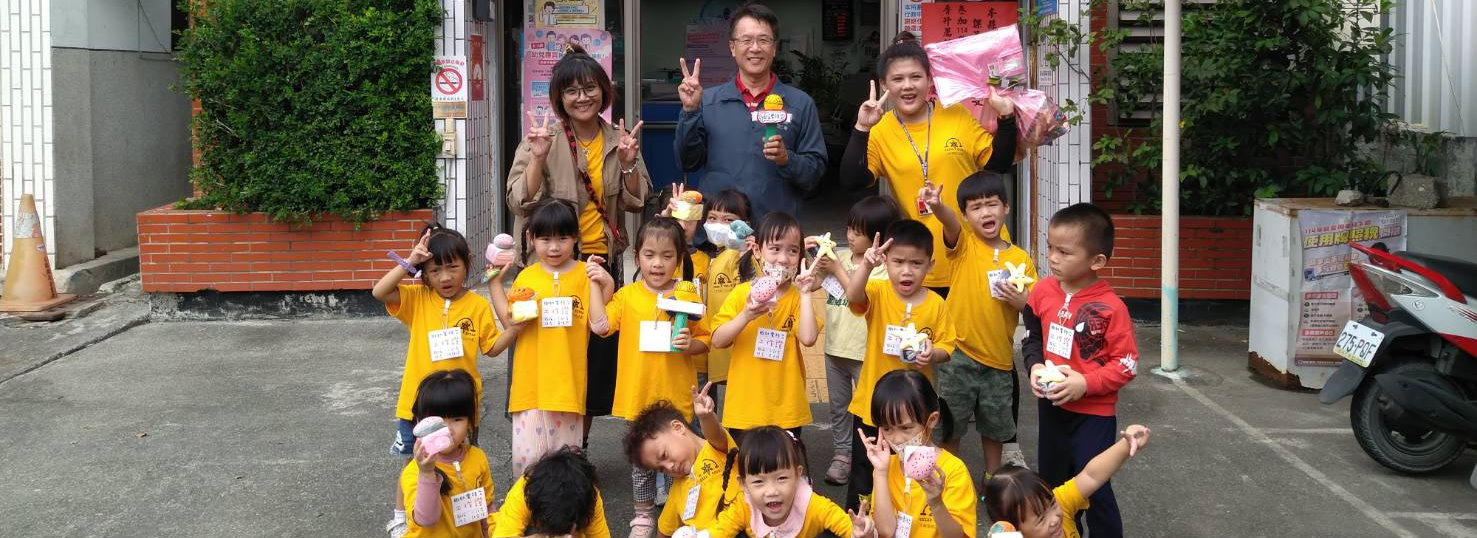 這張照片拍攝於戶政事務所門口，畫面中有約十五位穿著黃色制服的小朋友，孩子戴著名牌，站成兩排或蹲坐於前排。他們手中拿著彩色手工藝品(麥克風)，臉上露出愉快的神情，氣氛活潑。後方站立著幾位成人，包括幼兒園老師以及一位身穿深色服裝的戶政事務所秘書，大家一同比出和平手勢。背景可見辦公室入口、告示牌與盆栽。這是太麻里鄉立幼兒園主題課程「小小記者」活動，小朋友們訪問戶政秘書，獲得詳細解答，展現熱烈交流與學習精神。